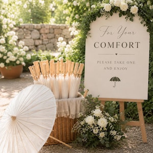 Peut inclure: Un présentoir de parapluies en papier blanc avec des manches en bois dans un panier en osier, à côté d'une pancarte indiquant "For Your Comfort". La pancarte est ornée de roses blanches et de verdure. Un parapluie ouvert est posé au sol.