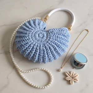 Peut inclure: Un sac à main en forme de coquillage crocheté bleu clair avec une poignée en corde blanche et des ferrures dorées. Un collier de perles, un collier doré avec un pendentif perle, un miroir compact et des coquillages sont également présents.