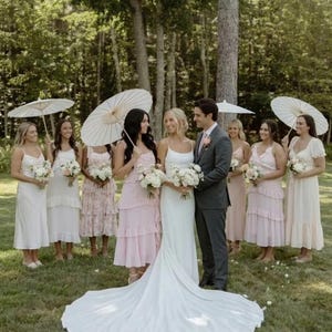 Peut inclure: Scène de mariage avec une mariée en robe blanche et un marié en costume gris, entourés de demoiselles d'honneur en robes roses et blanches. Les demoiselles d'honneur tiennent des bouquets et des parasols. Le cadre est un espace herbeux avec des arbres.
