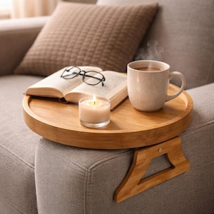 Puede incluir: Una bandeja redonda de bambú para el brazo del sofá con una vela encendida, una taza humeante, un libro abierto y gafas. La bandeja tiene un accesorio de reposabrazos de madera. Una almohada marrón descansa en el fondo.