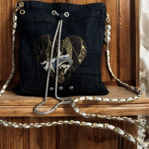 May include: A dark denim crossbody bag with a heart-shaped patch featuring a koi fish design. The bag has a drawstring closure and a silver chain strap with white and gold accents. Decorative pearls hang from the strap.