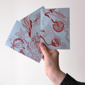 May include: Three light blue greeting cards with red floral designs are held in a hand. The cards feature intricate illustrations of flowers and botanical elements. The text "Congrats!" is visible on one card.