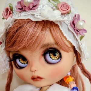 Può includere: Una bambola con capelli rosa, grandi occhi marroni e una cuffia in pizzo bianco decorata con fiori in tessuto rosa e bianchi. La bambola indossa un vestito color pesca con colletto bianco, bottoni di perla e un accessorio di perline con la scritta ANOUK.