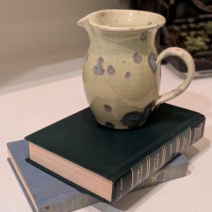 Puede incluir: Jarra de cerámica hecha a mano con asa y pico, colocada sobre dos libros. La jarra es de color verde claro con manchas azul oscuro y morado. Los libros son de color verde oscuro y azul.