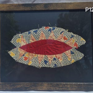 May include: Framed vintage textile art. The artwork features a central red velvet shape, surrounded by a patterned fabric with blue, yellow, and red heart and floral motifs. The piece is set against a black background within a wooden frame.