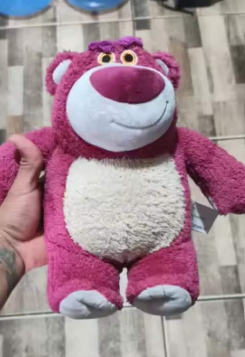 May include: A plush, pink bear with a white belly, a large pink nose, and small, yellow eyes. The bear has a friendly smile and is holding a tag. The bear's feet are light blue. This stuffed animal is a popular character.