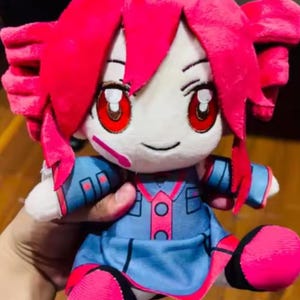 May include: A plush doll with a cartoon-like design. It features bright pink hair and accents, red eyes, and a blue outfit with pink details. The doll is soft and cuddly, with a smiling expression.