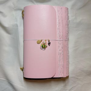 May include: A pink journal with a decorative lace edge and gold-coloured corner accents. The journal is secured with a white elastic band and features three decorative charms. The journal is closed.