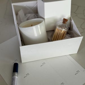 May include: A white gift box contains a white candle in a ceramic container, a small white box, and a glass bottle of matches. The candle has a wooden wick. Labels and a blue marker are also visible.