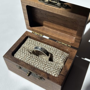 Puede incluir: Un anillo de plata con detalles grabados descansa dentro de un joyero de madera oscura abierto. La caja está forrada con una tela beige y tiene un cierre de latón. El anillo es el punto focal, lo que sugiere un tema de joyería o regalo.