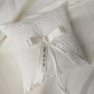 May include: A square white ring bearer pillow featuring an intricate floral lace overlay. A white satin ribbon bow is centered on the pillow, accented with small faux pearls. The ribbons are printed with the text Mr. & Mrs. Farkas and 24th June 2026.