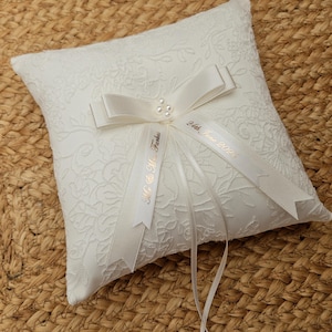 May include: A square white ring pillow featuring a textured floral lace overlay. A central white satin ribbon bow is accented with three small pearls. Two trailing ribbons are printed with the text Mr. & Mrs. Farkas and 24th, June 2025.