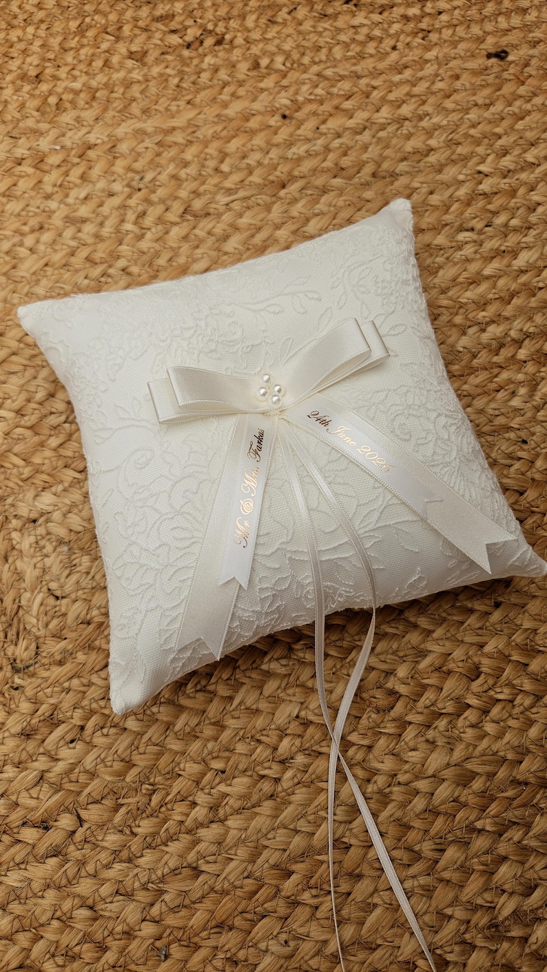 May include: A square white ring pillow featuring a textured floral lace overlay. A central white satin ribbon bow is accented with three small pearls. Two trailing ribbons are printed with the text Mr. & Mrs. Farkas and 24th, June 2025.