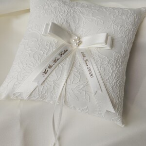 May include: A square white ring pillow featuring a textured floral lace overlay. A satin ribbon bow is centered on the pillow, accented with small white pearls. Two ribbon tails are printed with the text Mr. & Mrs. Farkas and 24th June 2026.