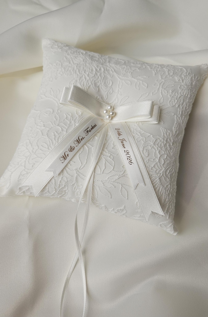 May include: A square white ring pillow featuring a textured floral lace overlay. A satin ribbon bow is centered on the pillow, accented with small white pearls. Two ribbon tails are printed with the text Mr. & Mrs. Farkas and 24th June 2026.