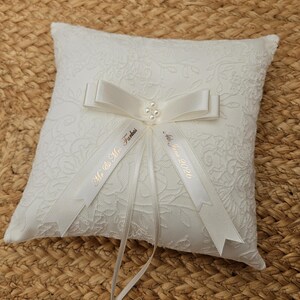 May include: A square white ring bearer pillow featuring a textured floral lace overlay. A satin ribbon bow is centered on the pillow, accented with four small white pearls. Two ribbon tails are printed with the text Mr. & Mrs. Farkas and 4th June 2026.