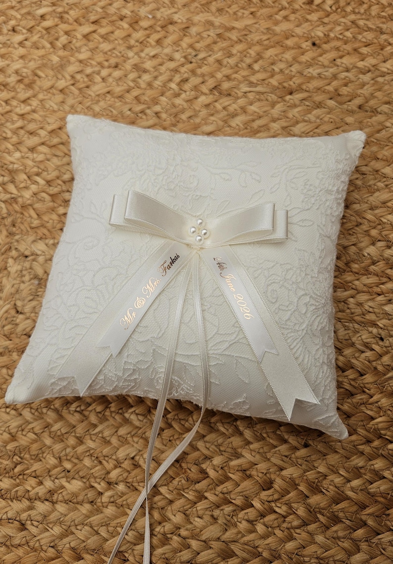 May include: A square white ring bearer pillow featuring a textured floral lace overlay. A satin ribbon bow is centered on the pillow, accented with four small white pearls. Two ribbon tails are printed with the text Mr. & Mrs. Farkas and 4th June 2026.