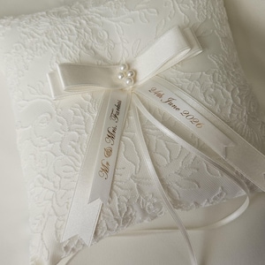 May include: A square ivory ring pillow featuring a delicate floral lace overlay. A satin ribbon bow is centered with a cluster of four small faux pearls. Two satin ribbons extend from the center, printed with the text Mr. & Mrs. Farkas and 24th June 2026.