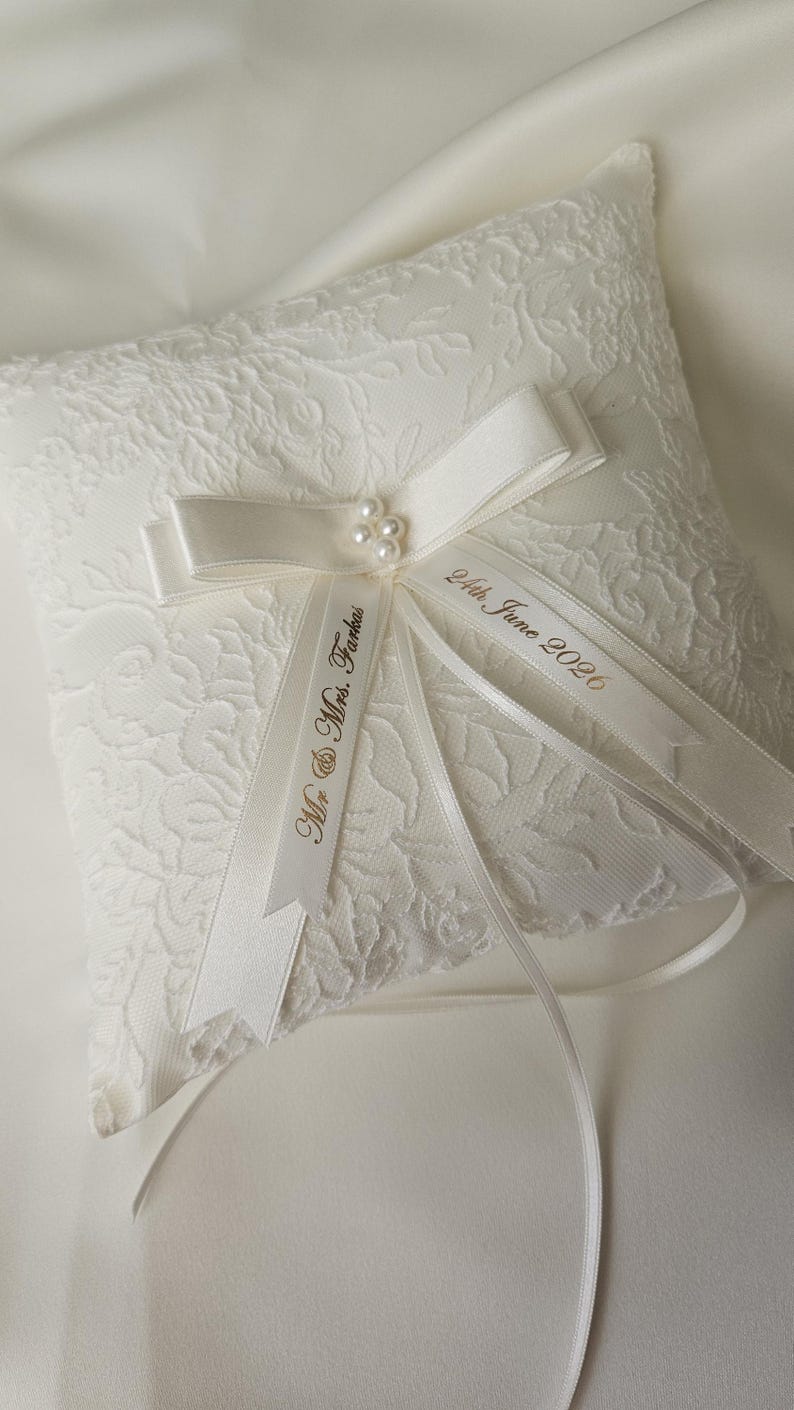 May include: A square ivory ring pillow featuring a delicate floral lace overlay. A satin ribbon bow is centered with a cluster of four small faux pearls. Two satin ribbons extend from the center, printed with the text Mr. & Mrs. Farkas and 24th June 2026.