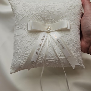 May include: A square white ring pillow featuring an intricate floral lace overlay. The center is adorned with a satin ribbon bow, four small faux pearls, and two trailing ribbons printed with the text Mr. & Mrs. Farkas and 24th June 2026.