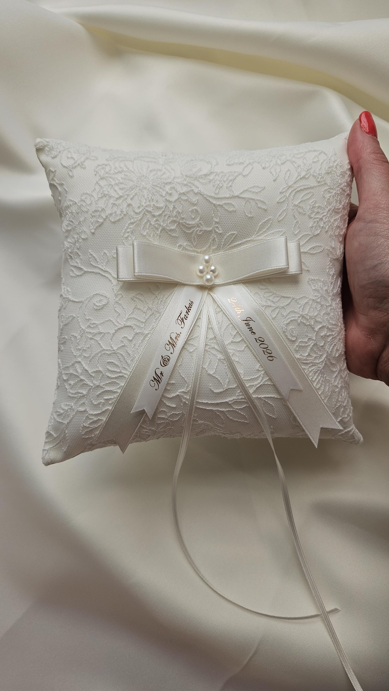 May include: A square white ring pillow featuring an intricate floral lace overlay. The center is adorned with a satin ribbon bow, four small faux pearls, and two trailing ribbons printed with the text Mr. & Mrs. Farkas and 24th June 2026.