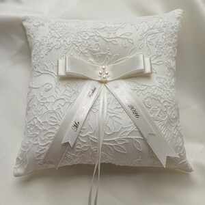 May include: A square white ring pillow featuring an intricate floral lace overlay. A satin ribbon bow with a small cluster of faux pearls sits at the center. Two hanging ribbons are printed with the text Mr. & Mrs. Farkas and 24th June 2026.