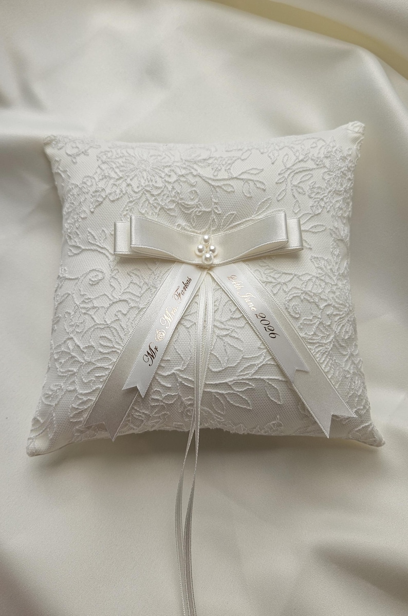 May include: A square white ring pillow featuring an intricate floral lace overlay. A satin ribbon bow with a small cluster of faux pearls sits at the center. Two hanging ribbons are printed with the text Mr. & Mrs. Farkas and 24th June 2026.