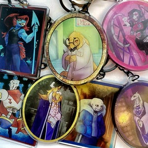May include: A collection of six acrylic keychains featuring various illustrated characters. The designs include a blue fish-like figure with a spear, a yellow reptilian scientist, a pink robotic character, a skeleton in armour, a king, and a goat-like figure.