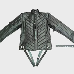 Op de afbeelding: Een zwarte jas met een gewatteerd chevronpatroon, een hoge kraag met gesp en lange mouwen met geïntegreerde riemen. Het kledingstuk bevat meerdere verstelbare lederachtige banden met metalen gespen en D-ringen op de borst, taille en mouwen.