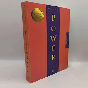 Puede incluir: Un libro de tapa dura titulado "The 48 Laws of Power" con una cubierta roja y una franja vertical púrpura. La palabra "POWER" está en letras doradas grandes. Una pegatina dorada de "National Bestseller" está en la portada.