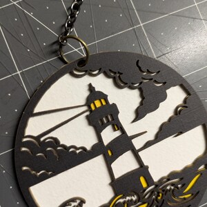 May include: A round, layered wooden ornament featuring a lighthouse scene. The lighthouse is black and white with yellow accents, set against a white background with black waves and clouds. A dark chain is attached.