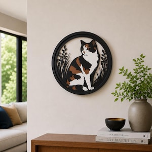 May include: A round, black cat wall art piece. The cat is depicted in a sitting position with a calico pattern of white, brown, and black. The cat is surrounded by black grass and framed by a black circular border. The art is mounted on a light beige wall.