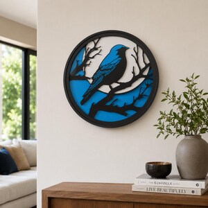 Puede incluir: Una obra de arte mural redonda y en capas que presenta un pájaro azul posado en una rama dentro de un marco circular negro. El fondo tiene una combinación de colores azul y blanco. La obra de arte está montada en una pared blanca.