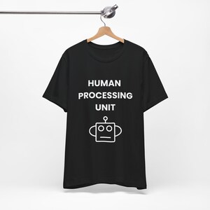 Puede incluir: Camiseta negra con el texto blanco "HUMAN PROCESSING UNIT" y un dibujo de un robot. La camiseta está colgada en una percha de madera sobre un fondo blanco.