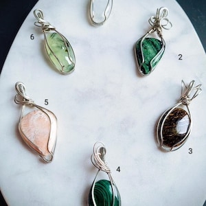 May include: Six gemstone pendants with silver wire wrapping are displayed on a white marble surface. The pendants feature various stones, including a moonstone, malachite, and a green stone. The pendants are numbered 1 through 6.