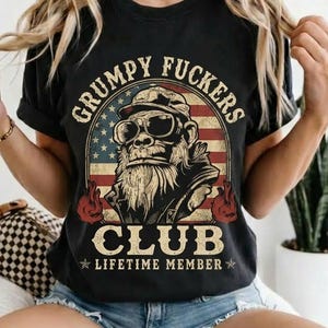 May include: Black t-shirt with the text "GRUMPY FUCKERS CLUB LIFETIME MEMBER" and a graphic of a gorilla wearing sunglasses and a hat, set against an American flag design. The shirt is a crew neck style.