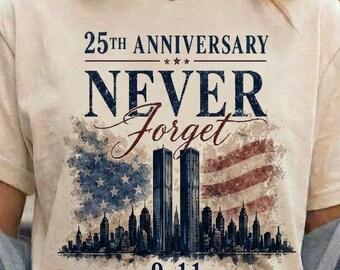 May include: A cream-coloured short-sleeve t-shirt featuring a graphic print with the text 25TH ANNIVERSARY NEVER FORGET 9.11 REMEMBER HONOR UNITE. The design includes a watercolour-style American flag and a silhouette of the Twin Towers skyline.