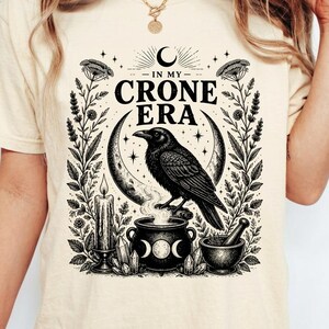 May include: Cream-colored t-shirt with a black graphic featuring a raven perched on a crescent moon, above a cauldron. The text "IN MY CRONE ERA" is displayed. Additional elements include a candle, herbs, and a mortar and pestle.