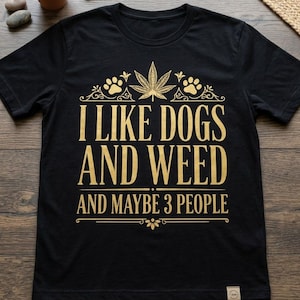 Può includere: T-shirt nero con testo dorato: "I LIKE DOGS AND WEED AND MAYBE 3 PEOPLE." Il design include una foglia di cannabis, impronte di zampe e decorazioni. La maglietta è realizzata in materiale morbido.