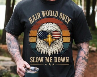 May include: A black short-sleeve t-shirt featuring a graphic of a bald eagle wearing sunglasses with an American flag pattern. The design includes a sunset background and the text "HAIR WOULD ONLY SLOW ME DOWN" in a vintage-style font.
