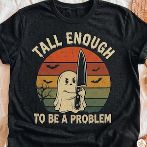 Puede incluir: Una camiseta negra de manga corta con un gráfico de un fantasma sosteniendo un cuchillo grande. El diseño incluye un fondo de atardecer con siluetas de murciélagos y un árbol, y el texto "TALL ENOUGH TO BE A PROBLEM" en una fuente desgastada.