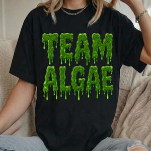Peut inclure: Un t-shirt noir à manches courtes avec le texte TEAM ALGAE imprimé dans une police vert vif, dégoulinante et semblable à de la vase. Le motif est centré sur la poitrine du t-shirt, qui possède une encolure ronde et une coupe standard.