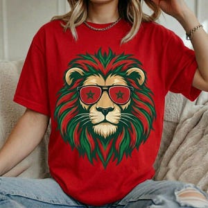 May include: A red short-sleeved t-shirt featuring a graphic of a lion's face with a green and red mane. The lion is wearing black-rimmed sunglasses with a green star on each lens. The shirt has a crew neckline and a relaxed fit.