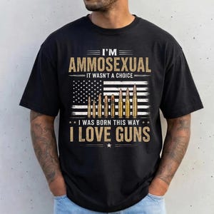 Può includere: Una t-shirt nera a maniche corte con una stampa grafica che riporta il testo I'M AMMOSEXUAL IT WASN'T A CHOICE I WAS BORN THIS WAY I LOVE GUNS. Il design include una bandiera americana stilizzata con illustrazioni di proiettili al posto delle strisce.