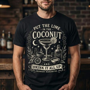 May include: A black t-shirt featuring a white graphic illustration of a margarita glass, a lime slice, and bottles. The text reads: PUT THE LIME IN THE COCONUT AND DRINK IT ALL UP MIDNIGHT MARGARITAS. The design includes stars, a crescent moon, and leaves.