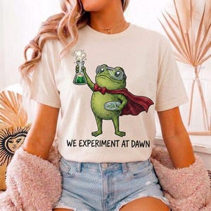 May include: A cream-colored t-shirt featuring a graphic of a green frog wearing goggles and a red cape. The frog holds a bubbling green flask and a petri dish. Below the illustration, the text reads, WE EXPERIMENT AT DAWN in black letters.