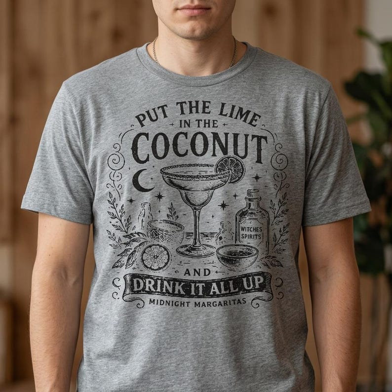 May include: A heather grey t-shirt featuring a black graphic print of a margarita glass, a lime slice, and a bottle labelled Witches Spirits. The text reads Put the Lime in the Coconut and Drink it all up, with Midnight Margaritas at the bottom.