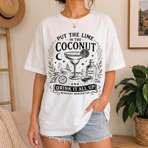 May include: A white short-sleeve t-shirt featuring a black graphic print of a margarita glass, a lime slice, and a bottle labelled Witches Spirits. The text reads Put the lime in the coconut and drink it all up with Midnight Margaritas below.