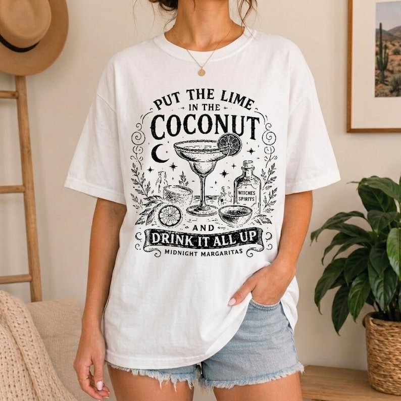 May include: A white short-sleeve t-shirt featuring a black graphic print of a margarita glass, a lime slice, and a bottle labelled Witches Spirits. The text reads Put the lime in the coconut and drink it all up with Midnight Margaritas below.