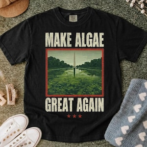 Könnte beinhalten: Ein schwarzes Kurzarm-T-Shirt mit dem Text "MAKE ALGAE GREAT AGAIN" in verwaschener weißer Schrift. Die mittlere Grafik zeigt einen grünen Teich mit Seerosen und das Washington Monument in der Ferne, umrahmt von einem roten Rand mit drei roten Sternen.